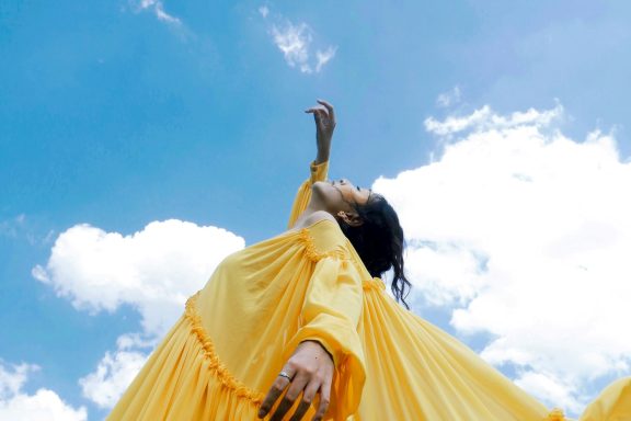 Frau in gelbem Kleid streckt die Arme in den blauen Himmel mit Wolken.