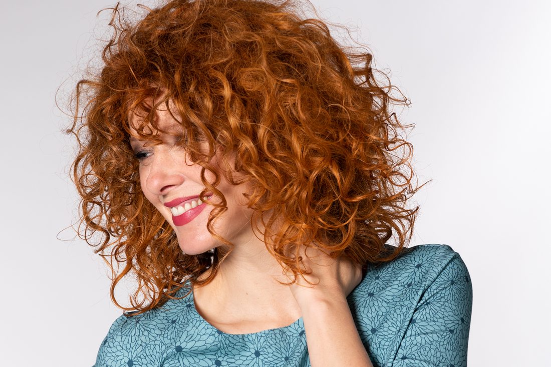 Frau mit lockigen, roten Haaren in blauer, gepunkteter Bluse, lächelt.